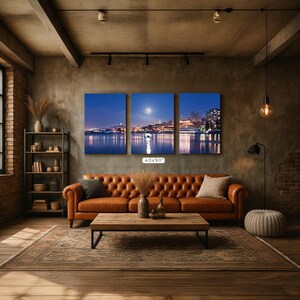 A cozy living room with a large, three-panel landscape painting depicting a city skyline at night, featuring a full moon and a river reflecting the city lights. The room has a rustic, industrial feel with exposed brick walls, a wooden floor, and a tufted ottoman.