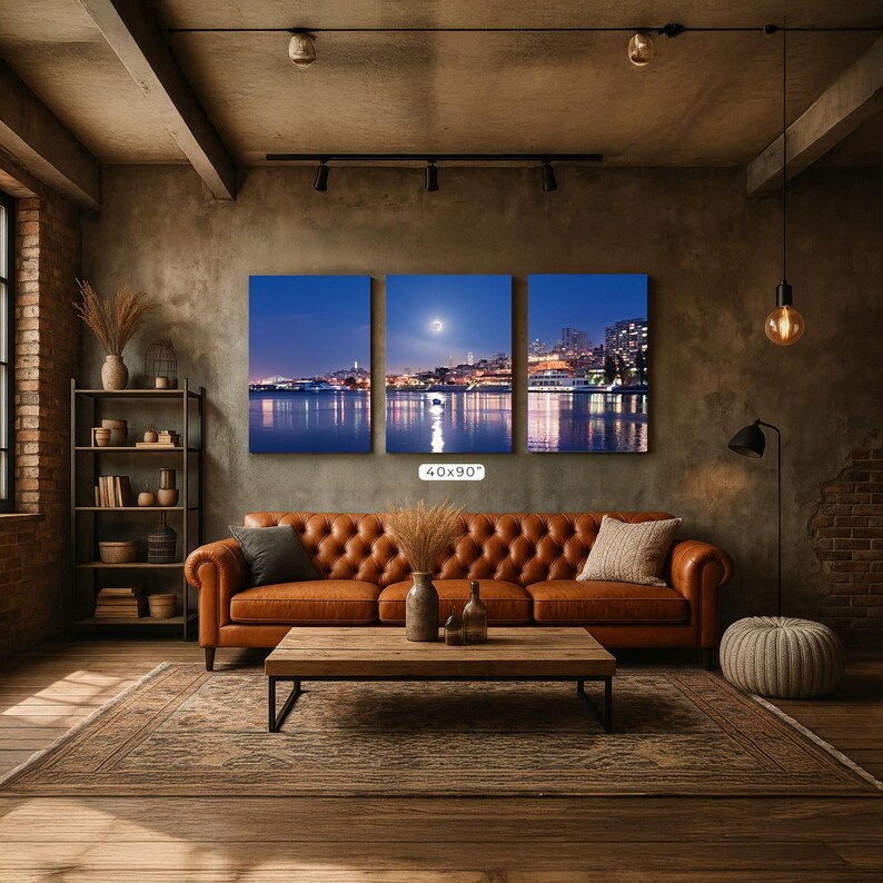 A cozy living room with a large, three-panel landscape painting depicting a city skyline at night, featuring a full moon and a river reflecting the city lights. The room has a rustic, industrial feel with exposed brick walls, a wooden floor, and a tufted ottoman.