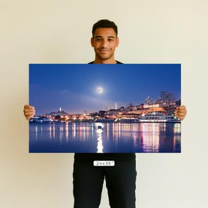 A man is holding up a large framed print of a nighttime cityscape with a crescent moon in the sky.