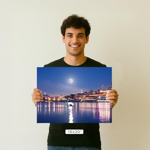 A young man is holding up a framed photograph of a cityscape at night, with a full moon visible in the sky.