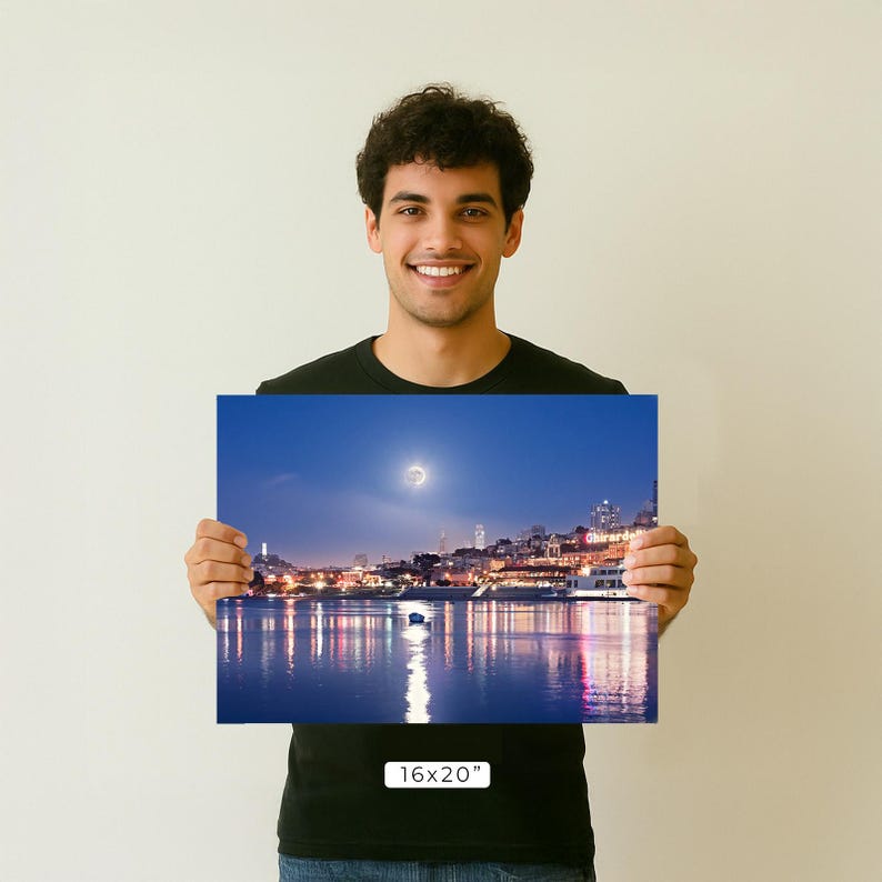 A young man is holding up a framed photograph of a cityscape at night, with a full moon visible in the sky.