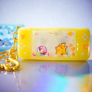Peut inclure: Un porte-clés en résine jaune en forme de console de jeu portable rétro. Le porte-clés présente des graphismes pixélisés d'un personnage, d'une balle et d'étoiles. La quincaillerie dorée relie le porte-clés à un porte-clés.