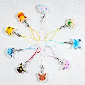 Peut inclure: Une collection de breloques de personnages colorés avec des fermoirs argentés et des lanières de poignet assorties. Les breloques présentent divers personnages de dessins animés dans des tons de jaune, orange, bleu, rose et marron. Les breloques sont disposées en cercle.