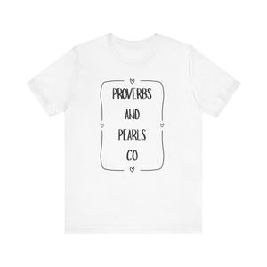 May include: White t-shirt with the text "PROVERBS AND PEARLS CO" in a black, outlined rectangular shape. The text is centered on the shirt, with small heart shapes at each corner of the rectangle. The shirt has short sleeves and a crew neck.