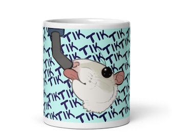 May include: White ceramic mug with a cartoon rat on a light blue background. The rat is sticking its tongue out and has a black eye. The background is covered in the word "Tik" repeated in a dark blue color.