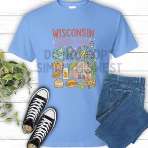 May include: Pale blue t-shirt featuring a vibrant graphic advertising the Wisconsin State Fair. The design includes a Ferris wheel, food items, and the text "WISCONSIN State Fair". Black and white trainers and blue jeans are also in the image.