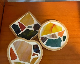Può includere: Tre sottobicchieri a mosaico di forme diverse: un quadrato, un cerchio e un cuore. Ogni sottobicchiere presenta una cornice in legno e un colorato disegno a mosaico con sfumature di giallo, arancione, verde e rosso. I sottobicchieri sono esposti su una superficie di legno.