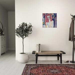 May include: An abstract painting of a woman with pink hair and closed eyes. The artwork features a colorful background with blue, yellow, and purple hues. The painting is displayed on a white wall in a well-lit room.