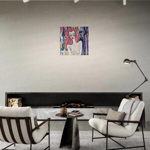 May include: An abstract painting of a woman with red hair and a nude torso. The artwork is displayed on a white wall in a modern living room setting. The painting features a colorful background with vertical brushstrokes.