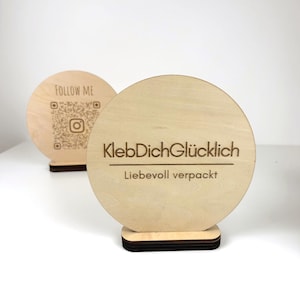 Könnte beinhalten: Zwei runde Schilder aus hellem Holz. Eines zeigt "FOLLOW ME" und einen QR-Code. Das andere Schild trägt die Aufschrift "KlebDichGlücklich" und "Liebevoll verpackt". Beide Schilder sind auf einem dunkelbraunen Sockel montiert.