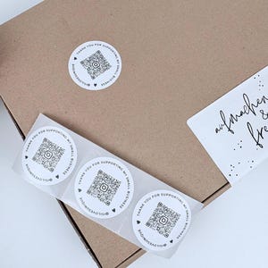 Könnte beinhalten: Ein brauner Karton mit mehreren weißen Aufklebern. Die Aufkleber haben einen QR-Code, den Text "THANK YOU FOR SUPPORTING MY SMALL BUSINESS" und den Instagram-Handle "@ISILOVESUNIQUE". Ein weißes Etikett mit handschriftlichem Text ist ebenfalls sichtbar.