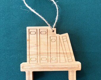 Puede incluir: Adorno de estantería de madera con una cuerda para colgar. El adorno presenta una estantería con cuatro libros, uno de los cuales está inclinado. La madera tiene un acabado natural y es adecuado para un árbol de Navidad u otra decoración navideña.