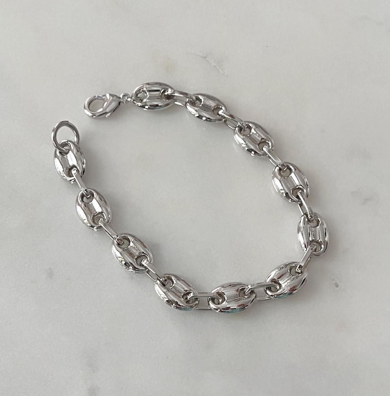 May include: A silver-colored chain bracelet with a lobster clasp. The bracelet features a series of oval-shaped links, creating a bold and modern design. The links are connected to form a flexible chain, suitable for everyday wear.