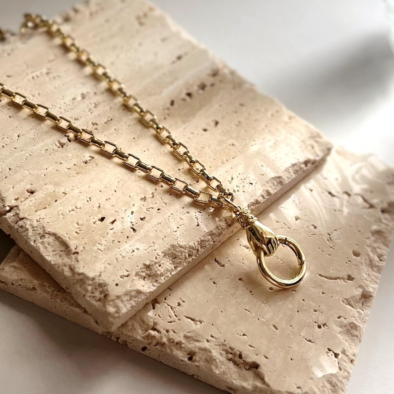 May include: A gold-toned chain necklace featuring a clasp shaped like a hand holding a circular ring. The chain consists of uniform rectangular links connected in a series, with the hand-shaped clasp serving as the focal point at the end.