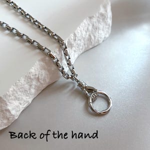 May include: A silver-toned chain necklace featuring a pendant shaped like a hand holding a circular ring. The pendant depicts the back of the hand with detailed knuckles and wrist texture. The chain consists of rectangular links in a polished finish.