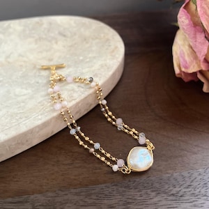 May include: A delicate gold bracelet featuring two strands of small, gold beads and interspersed pink and gray gemstones. A large, iridescent, oval-shaped stone is set in a gold wire frame. The bracelet has a gold toggle clasp.