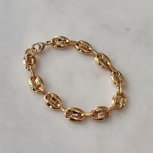 May include: A gold-colored bracelet featuring a series of oval-shaped links, creating a chain-like design. The bracelet is made of a shiny, metallic material and has a clasp closure. The jewelry is displayed on a light-colored surface.