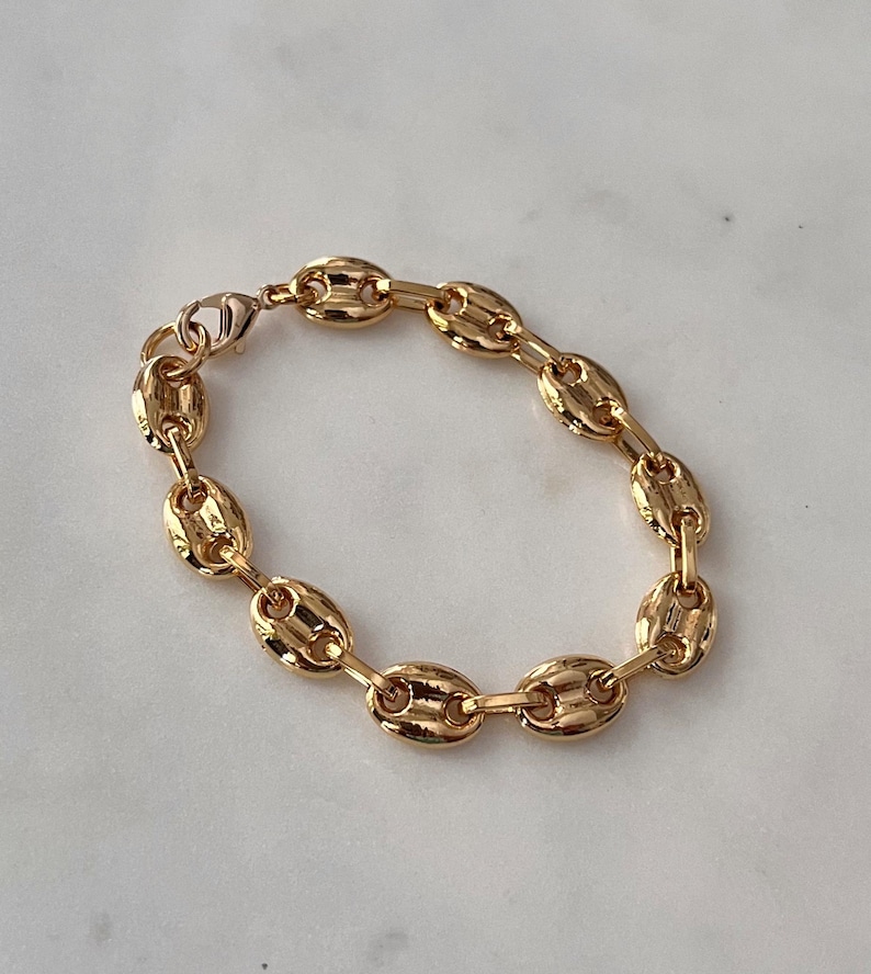 May include: A gold-colored bracelet featuring a series of oval-shaped links, creating a chain-like design. The bracelet is made of a shiny, metallic material and has a clasp closure. The jewelry is displayed on a light-colored surface.