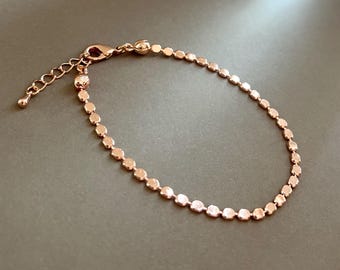 May include: Rose gold bracelet featuring a delicate chain of small, flat, circular links. The bracelet has an adjustable length and a lobster clasp closure. The jewelry is displayed on a neutral gray background.