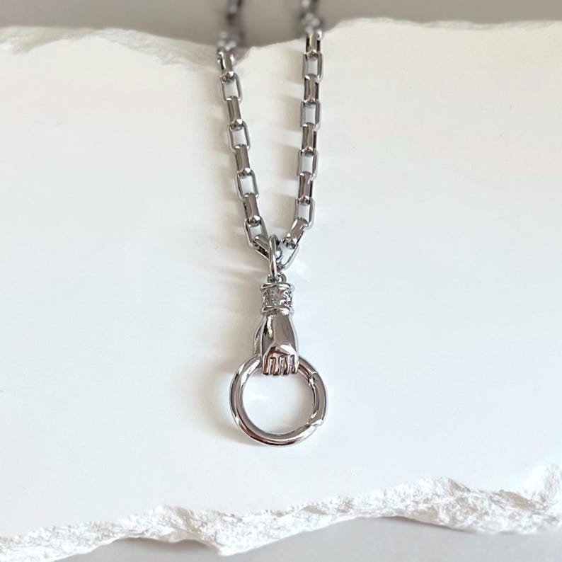 May include: A silver-toned necklace featuring a chain with rectangular links and a pendant shaped like a hand holding a circular ring. The metallic finish is polished and reflective, showcasing a detailed hand design grasping the bottom loop.