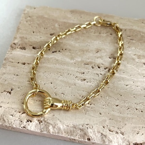 May include: A gold-toned chain bracelet featuring a series of rectangular links and a circular pendant. The pendant is held by a sculpted hand motif with intricate detailing at the wrist, connecting the circular ring to the main chain structure.