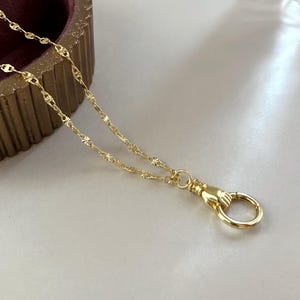 May include: A gold-toned necklace featuring a delicate chain with small, star-like textured links. The chain terminates in a functional clasp shaped like a hand holding a circular ring, providing a secure closure for this piece of jewelry.