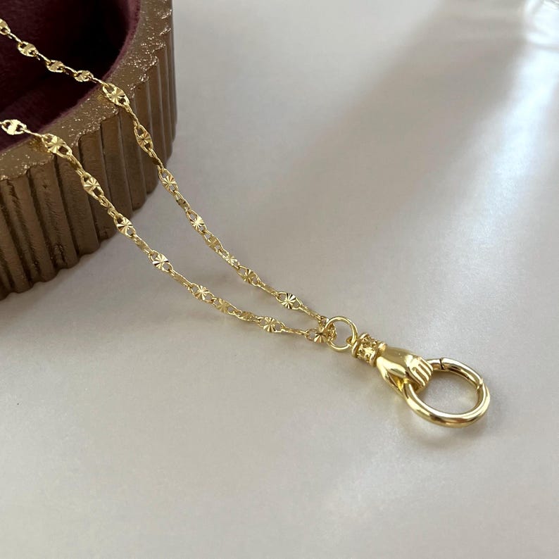 May include: A gold-toned necklace featuring a delicate chain with small, star-like textured links. The chain terminates in a functional clasp shaped like a hand holding a circular ring, providing a secure closure for this piece of jewelry.