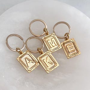 Op de afbeelding: Vier goudkleurige sleutelhangers met rechthoekige hangers. Elke hanger heeft een andere letter: A, K, M en E. De letters zijn in een schreefletter geëmbosseerd. De sleutelhangers hebben een eenvoudig, elegant ontwerp.