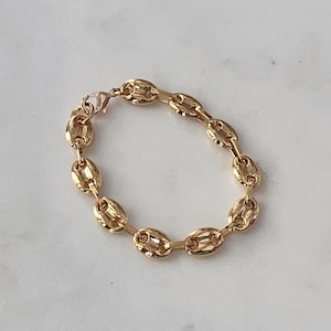 May include: A gold-colored bracelet featuring a series of oval-shaped links. The bracelet is made of a shiny, metallic material and is fastened with a lobster clasp. The jewelry is displayed on a light-colored surface.