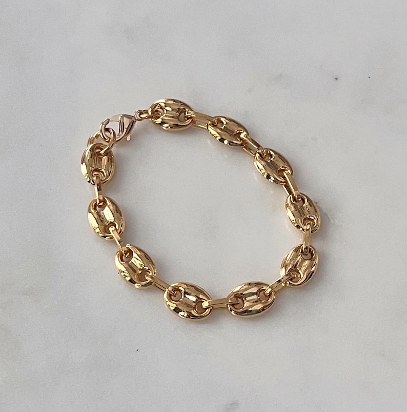 May include: A gold-colored bracelet featuring a series of oval-shaped links. The bracelet is made of a shiny, metallic material and is fastened with a lobster clasp. The jewelry is displayed on a light-colored surface.
