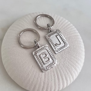 Può includere: Due portachiavi argentati con ciondoli rettangolari. Un ciondolo presenta la lettera "B", l'altro "J". Ogni ciondolo ha un bordo decorativo ed è attaccato a un portachiavi argentato. Esposti su una superficie bianca strutturata.