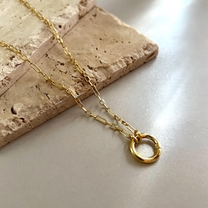 Peut inclure: Un collier doré composé d'une chaîne délicate aux maillons rectangulaires allongés. La chaîne est reliée à un pendentif unique en forme d'anneau circulaire lisse à la base, offrant un design simple et épuré avec une finition métallique polie.