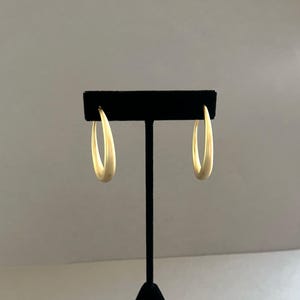 May include: A pair of gold-colored hoop earrings displayed on a black stand. The earrings have a smooth, matte finish and a teardrop shape. The background is a neutral gray.