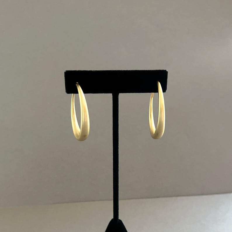 May include: A pair of gold-colored hoop earrings displayed on a black stand. The earrings have a smooth, matte finish and a teardrop shape. The background is a neutral gray.