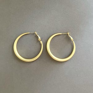 May include: A pair of gold-colored hoop earrings. The earrings have a matte finish and a simple, classic design. The hoops are round and have a hinged closure. The earrings are displayed on a gray background.