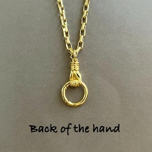 May include: A gold-toned necklace featuring a chain with rectangular links and a pendant shaped like a hand holding a circular ring. The pendant is positioned above the text "Back of the hand" which is printed in a simple black font.