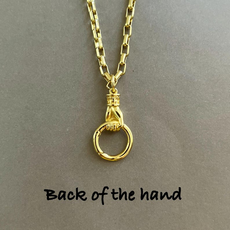 May include: A gold-toned necklace featuring a chain with rectangular links and a pendant shaped like a hand holding a circular ring. The pendant is positioned above the text "Back of the hand" which is printed in a simple black font.