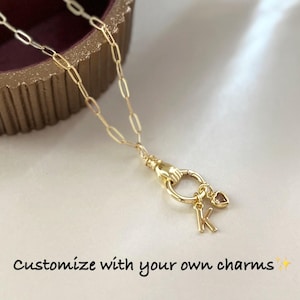May include: A gold-toned chain necklace featuring a clasp shaped like a hand holding a circular ring. Attached to the ring are a letter K charm and a small heart-shaped charm. The text "Customize with your own charms" is printed below.