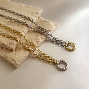May include: Two cable chain necklaces, one in a gold tone and one in a silver tone. Each necklace features a series of rectangular links and terminates in a circular ring clasp. The gold chain has a silver ring, while the silver chain has a gold ring.