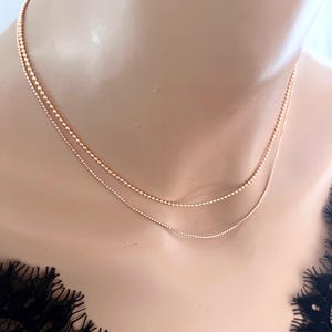 May include: A delicate, layered necklace with two strands of small, round beads. The necklace is a warm rose gold color and rests on a neutral background. The jewelry is a simple, elegant accessory.