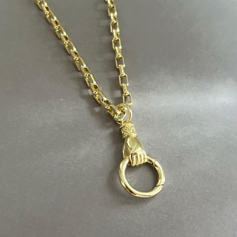 May include: A gold-toned chain necklace featuring a pendant shaped like a hand holding a circular ring. The chain consists of rectangular links, and the pendant depicts a detailed hand with a cuff at the wrist, securely grasping a smooth, round metal loop.