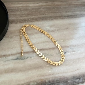 Könnte beinhalten: Ein goldfarbenes Armband mit einem sich wiederholenden Muster aus pfeilförmigen oder blattartigen Gliedern. Die Kette verfügt über einen Karabinerverschluss und eine verstellbare Verlängerungskette mit einem kleinen tropfenförmigen Anhänger.