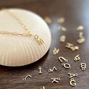 Puede incluir: Un collar dorado con un colgante con la letra "M". El collar está sobre una superficie blanca y redondeada, con otros abalorios de letras doradas esparcidos. El fondo es una superficie marrón oscuro.