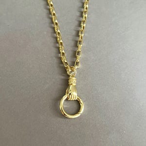 May include: A gold-toned necklace featuring a thick, rectangular link chain. The pendant is a sculpted hand holding a circular ring, with a decorative textured band at the wrist where the hand connects to the chain, creating a cohesive metallic piece.