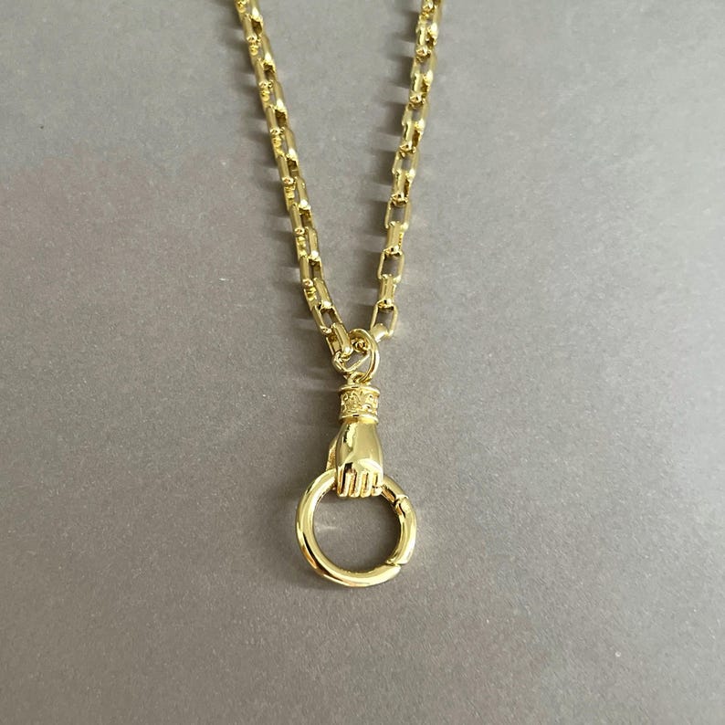 May include: A gold-toned necklace featuring a thick, rectangular link chain. The pendant is a sculpted hand holding a circular ring, with a decorative textured band at the wrist where the hand connects to the chain, creating a cohesive metallic piece.