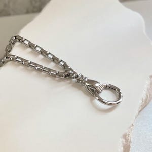 May include: A silver-toned box chain necklace featuring a unique clasp designed as a small hand holding a circular ring. The metallic links are square-shaped and polished, creating a reflective surface that catches the light along the entire length.