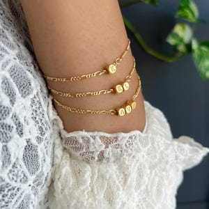 Puede incluir: Tres pulseras doradas con un delicado diseño de cadena. Cada pulsera presenta pequeñas cuentas cúbicas doradas con letras grabadas. Las letras de las cuentas deletrean "K", "KL" y "AVA".