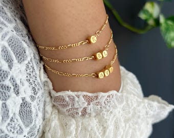 May include: Three gold-colored bracelets with a delicate chain design. Each bracelet features a series of small, gold-colored cube beads with engraved letters. The letters on the beads spell out "K", "KL", and "AVA".