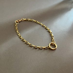May include: A gold-colored bracelet with a rectangular link chain design. The bracelet features a circular clasp and a lobster claw closure. The jewelry is displayed on a neutral gray surface.