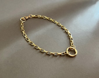 May include: A gold-colored bracelet with a rectangular link chain design. The bracelet features a circular clasp and a lobster claw closure. The jewelry is displayed on a neutral gray surface.